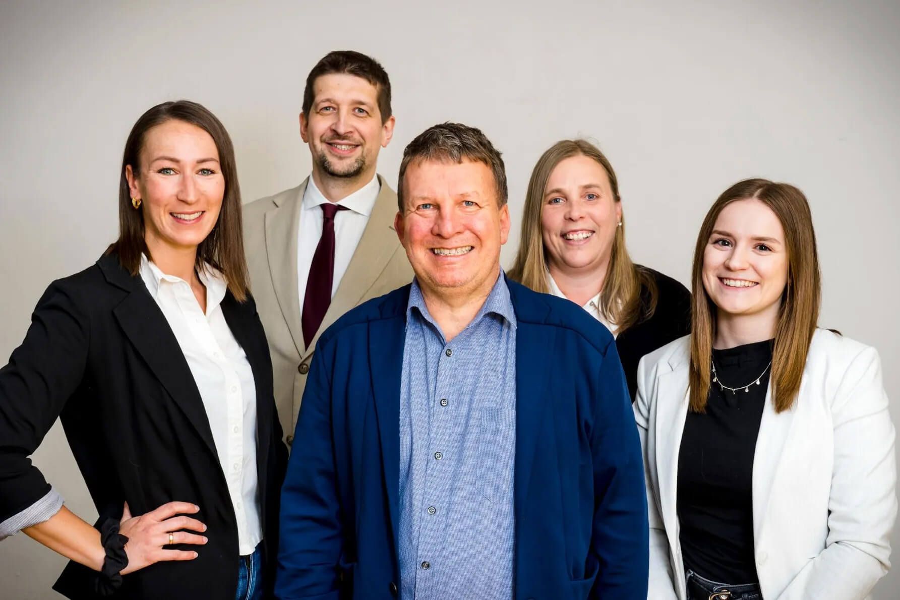 Fünf Personen stehen vor hellem Hintergrund, drei Frauen und zwei Männer, alle in Businesskleidung, die vordere Person trägt ein blaues Hemd und eine blaue Jacke.